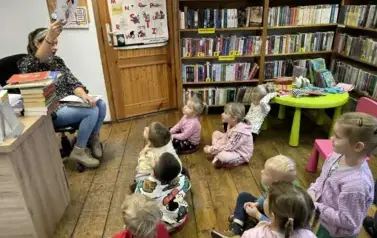 Dzieci siedzą na siedziskach i patrzą w kierunku bibliotekarki, kt&oacute;ra pokazuje im na kartkach bohater&oacute;w z bajki, a dzieci odgadują.