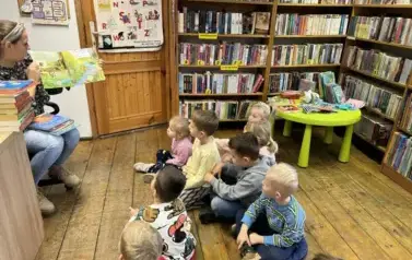 Dzieci siedzą na siedziskach w bibliotece i parzą na bibliotekarkę, kt&oacute;ra czyta im bajkę pokazując przy tym ilustracje.