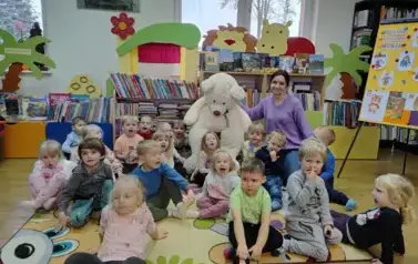 Wszystkie dzieci siedzą na dywanie, po środku na krzesełku siedzi duży biały miś, a obok siedzi bibliotekarka.