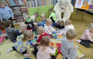 Dzieci siedzą na dywanie i bawią się pluszowymi książeczkami. Obok na krześle siedzi duży biały miś.