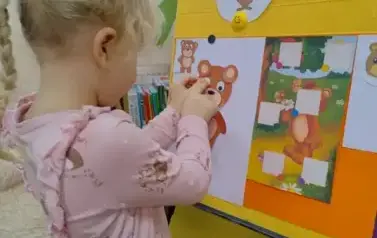 Dziewczynka w blond włosach w warkoczyku stoi przy tablicy i nakleja na kartkę części misia.