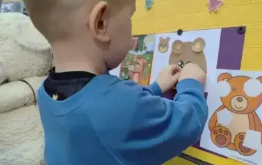 Chłopiec w blond włosach i w niebieskiej bluzie stoi przy tablicy i uzupełnia naklejając na kartkę części misia.