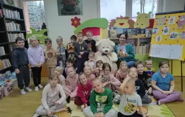 Wszystkie dzieci siedzą na dywanie, po środku na krzesełku siedzi duży biały miś, a obok siedzi bibliotekarka trzymając bajkę o niedźwiadku.