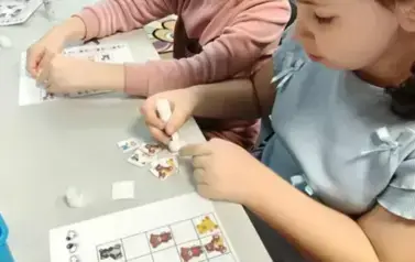 Dwie dziewczynki przy stole uzupełniają na kartach pracy misiowe sudoku naklejając w odpowiednie budki misiowe kwadraciki.