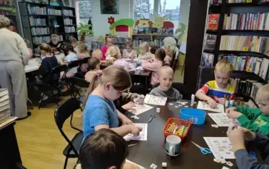 Trzy stoły pełne dzieci, kt&oacute;re uzupełniają miosiwe sudoku. Z lewej strony widać Panią, kt&oacute;ra pomaga przy stole. 