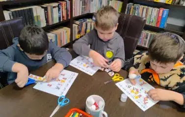 Troje chlopc&oacute;w przy stole naklejają w odpowiednie budki na kartach pracy kwadraciki z misiami  uzupełniając misiowe sudoku.