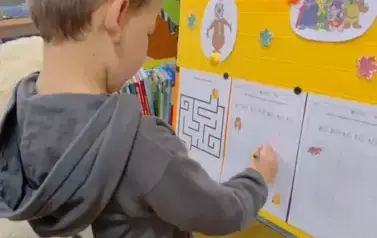 Przy tablicy przystrojonej misiowymi grafikami stoi chłopiec w szarej bluzie i uzupełnia kartę pracy prowadząc misia do miodu