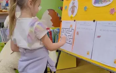 Przy tablicy przystrojonej misiowymi grafikami stoi dziewczynka w blond włosach w kitku i uzupełnia kartę pracy prowadząc misia do miodu