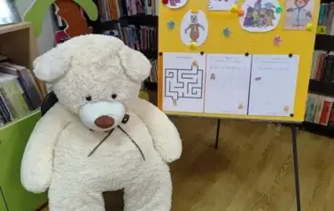 Po prawej stronie stoi tablica przystrojona grafikami misi&oacute;w i kartami pracy. Po lewej stronie tablicy siedzi na krześle duży biały miś. Za nim widać palmę kt&oacute;ra jest częścią szafki na ksiązki.