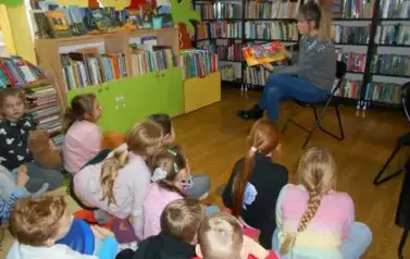 Dzieci siedzą na dywanie i patrzą na bibliotekarkę, kt&oacute;ra siedzi z przodu i czyta im bajkę mając skierowaną książkę z obrazkami w kieunku dzieci.