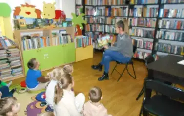 Dzieci siedzą na dywanie i patrzą na bibliotekarkę, kt&oacute;ra siedzi z przodu i czyta im bajkę mając skierowaną książkę z obrazkami w kieunku dzieci.