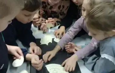 Dzieci stoją przy stole na kt&oacute;rym leży czarna kartka z wizerunkiem misia i dzieci naklejają na misia brakujące elementy wyklejanki.