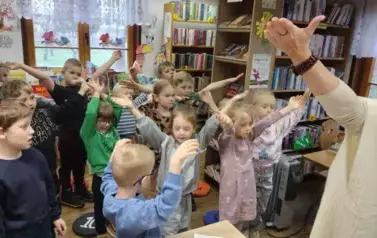 Dzieci stoją w rozsypce i mają uniesione ramiona do g&oacute;ry tak jak ich Pani prowadząca zajęcia. 