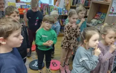 Dzieci stoją w rozsypce, niekt&oacute;re z nich mają położony palec na ustach jakby informowały innych o ciszy.