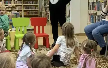 Dzieci siedzą na dywanie, a przed nimi stoi osoba przebrana w misia Pandę i się uśmiecha