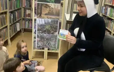 Dzieci siedzą na dywanie, a z przodu kobieta przebrana za misia Pandę pokazuje im bruszurę o segregowaniu śmieci.