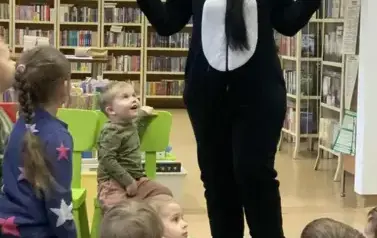 Dzieci siedzą na dywanie, a przy nich stoi kobieta przebrana za misia Pandę, ma rozłożone ręce w bok.