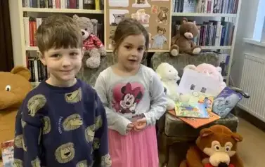 Przy fotelu z misiami stoi chłopiec i dziewczynka, obok na podłodze siedzi brązowy miś, a za nimi stoi korkowa tablica  z grafikami dot. święta misia.