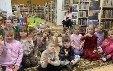 Dzieci siedzą na dywanie, z boku po prawej stonie na krześle siedzi Pani z Przedszkola.