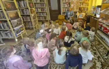 Dzieci siedzą na dywanie i patrzą do przodu na bibliotekarkę, kt&oacute;ra im opowaida o święcie Misia. Obok bibliotekarki siedzi duży brążowy miś, a po prawej na fotelu leżą małe misie i bajki do czytania. Na wprost stoi tablica przystrojna w grafikę o misiach