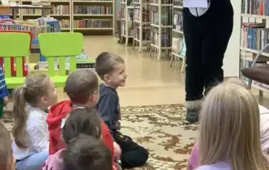 Dzieci siedzą na dywanie, a przed nimi stoi osoba przebrana w misia Pandę pochylona lekko ku dzieciom i trzymająca w rękach białe kartki.