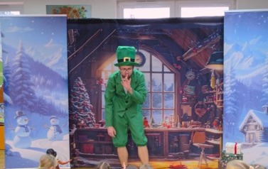 Po środku stoi pochylony elf i ma przyłożony palec do ust. W tle pok&oacute;j z choinką , a po bokach zaśnieżone choinki.