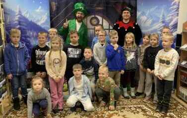 Na tle pokoju z choinką  i z bocznymi zaśnieżonymi choinkami stoją dzieci w grupie razem z dwoma elfami.