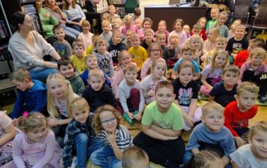 Na dywanie siedzi spora grupa dzieci, po bokach na krzesłach siedzą nauczyciele.