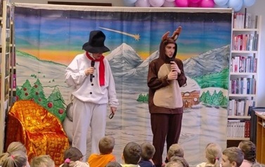 Na środku stoją przebrani bałwan i renifer i patrzą na dzieci. Obok stoi fotel ze złotoczerwonym nakryciem. W tle g&oacute;rski zaśnieżony pejzarz.