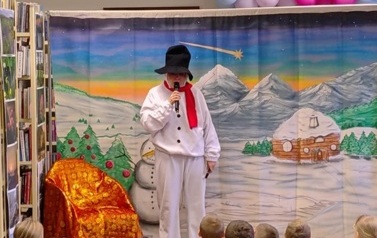 Na środku stoi przebrany bałwan i m&oacute;wi przez mikrofon. Obok stoi fotel ze złotoczerwonym nakryciem. W tle g&oacute;rski zaśnieżony pejzarz.