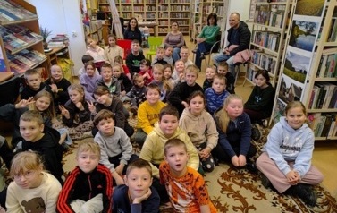 Dzieci siedzą liczną grupą na dywanie, za nimi na krzesłach siedzą opiekunowie.