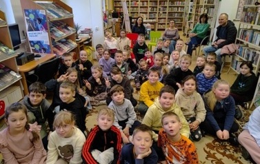 Dzieci siedzą liczną grupą na dywanie, za nimi na krzesłach siedzą opiekunowie.