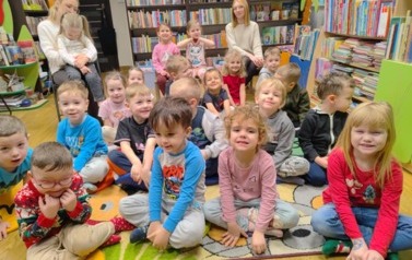 Grupa dzieci siedzi na dywanie, a za nimi z tyłu siedzi ich Pani, kt&oacute;ra na kolanach trzyma dziewczynkę, a obok na krzesełku siedzi druga ich Pani.