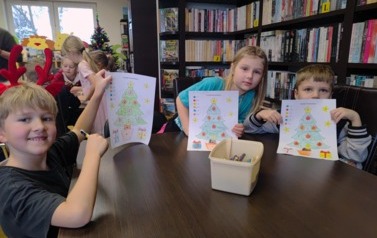 Dwoje chłopc&oacute;w i dziewczynka siedzą przy stole i pokazują swoje pokolorowane choinki.