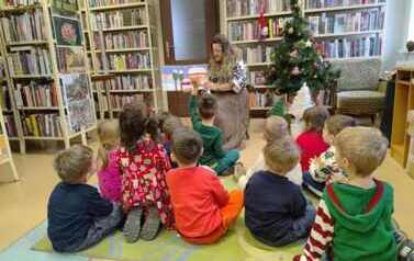 Dzieci siedzą na dywanie i patrzą na bibliotekarkę, kt&oacute;ra czyta bajkę z książeczki skierowanej obrazkami do dzieci.
