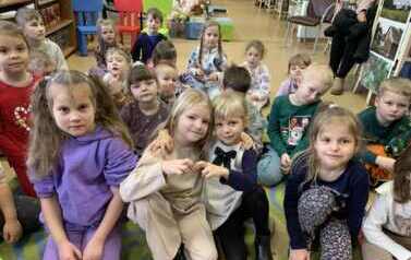 Dzieci siedzą razem na dywanie, dwie dziewczynki obejmują się razem i z rączek robią serduszko.