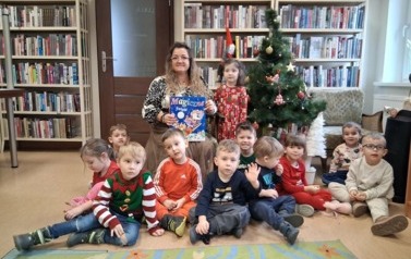 Dzieci siedzą przy choince z Bibliotekarką, ktora trzyma książeczkę pt "Magiczne Święta"