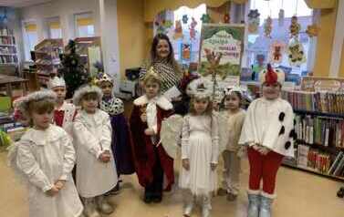 Uśmiechnięta bibliotekarka stoi razem z przedszkolnymi kolędnikami