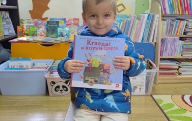Uśmiechnięty mały chłopiec siedzi na krzesełku i pokazuje nową książkę. 