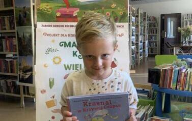 Chłopiec w blond włosach stoi z lekko wyciągniętą głową do przodu, trzyma książkę , kt&oacute;rą otrzymał. Za nim jest tablica korkowa z plakatem "Mała Książka Wielki Człowiek".