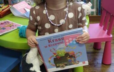 Uśmiechnięta dziewczynka w blond włosach do ramion trzyma swoją nową książeczkę, kartę młodego czytelnika i misia kr&oacute;wkę. Na szyi ma korale.