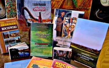 Książki regionalne wyłożone na stoliku, a za nimi obraz z tłem starej chaty kociewskiej