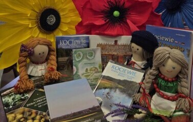 Książki regionalne wyłożone na stoliku z widocznymi tytułami obok laki kociewskie, a za nimi położone sztuczne korony kwiat&oacute;w w kolorze niebieskim, czerwonym i ż&oacute;łtym