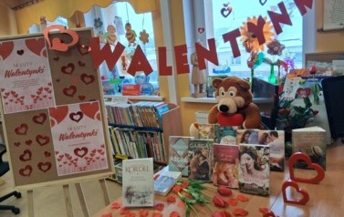 Na biurku wyłożone są książki o miłości, tulipany sztuczne i płatki kwiat&oacute;w, miś trzymający serduszko i drewniane serduszka, obok stoi tablica z plakatami walentynkowymi, a wyżej nad książkami wisi napis WALENTYNKI