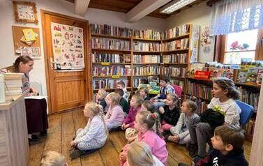 Dzieci siedzą na siedziskach i patrzą na bibliotekarkę, kt&oacute;ra im coś opowiada