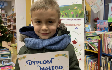 Uśmiechnięty chłopiec w blond włosach stoi w bibliotece w Zblewie trzymając sw&oacute;j dyplom. Za nim stoi tablica z plakatami projektu Mała Książka Wielki Człowiek