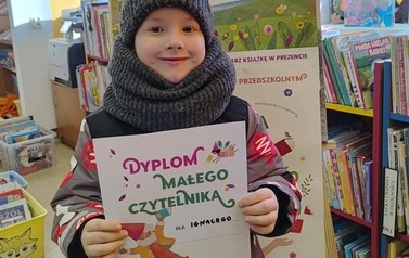 Uśmiechnięty chłopiec stoi w bibliotece w Zblewie trzymając sw&oacute;j dyplom. Za nim stoi korkowa tablica z plakatami projektu Mała Książka Wielki Człowiek.