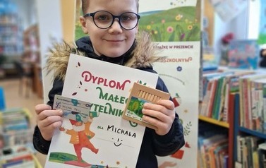 Chłopiec w ciemnych okularach i w blond włosach stoi w bibliotece w Zblewie pokazując sw&oacute;j dyplom, kartę Małego Czytalnika i kredki świecowe. Za nim stoi korkowa tablica z plakatami projektu Mała Książka Wielki Człowiek