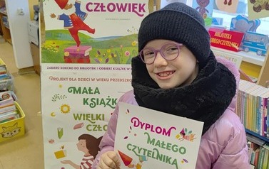 Uśmiechnięta dziewczynka w r&oacute;żowej kurtce i w r&oacute;zowych okularach, w czarnej czapce i szaliku pokazuje sw&oacute;j dyplom w bibliotece w Zblewie. Za nią stoi tablica z plakatami projektu Mała Książka Wielki Człowiek