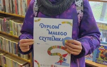 Dziewczynka w fioletowej kurtce stoi w bibliotece w Borzechowie i pokazuje sw&oacute;j dyplom.
